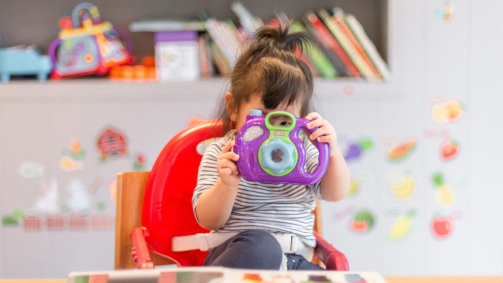 A young girl in a highchair peeks through the top of a toy camera.