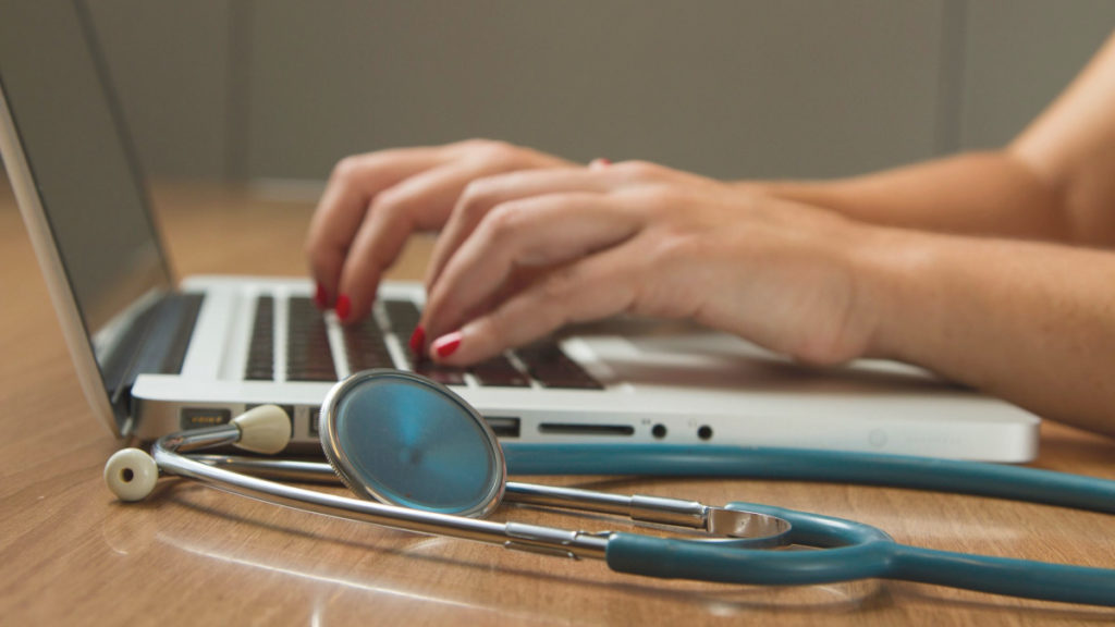 A person types on a laptop with a stethoscope next to them. Healthcare will be positively impacted by improved 5G coverage.