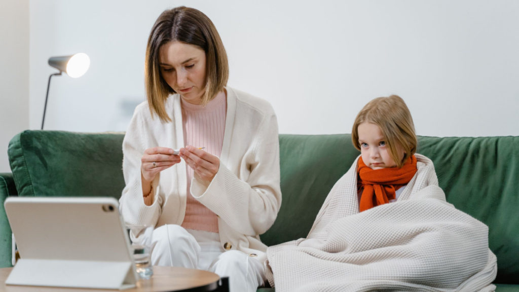 A woman sits beside a child on a couch, holding a thermometer. There is a tablet sitting on the table in front of them as they wait for a telemedicine appointment.