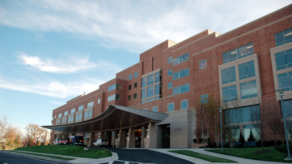 The outside of a rural hospital, showing the brick facade and vehicles lined up at the entrance.