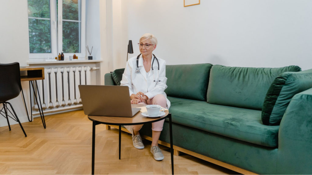 A telehealth provider sits on a green couch with a laptop sitting on a table in front of her.